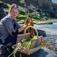 Oonagh on a small beach with rocks. She is holding a handful of seaweed and has a basket of foraged wild food beside her. Foraging in County Clare